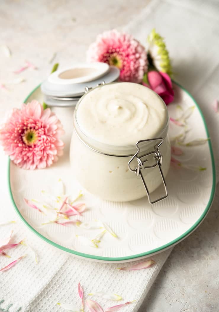 A glass jar of homemade lotion on a white plate with flowers around it