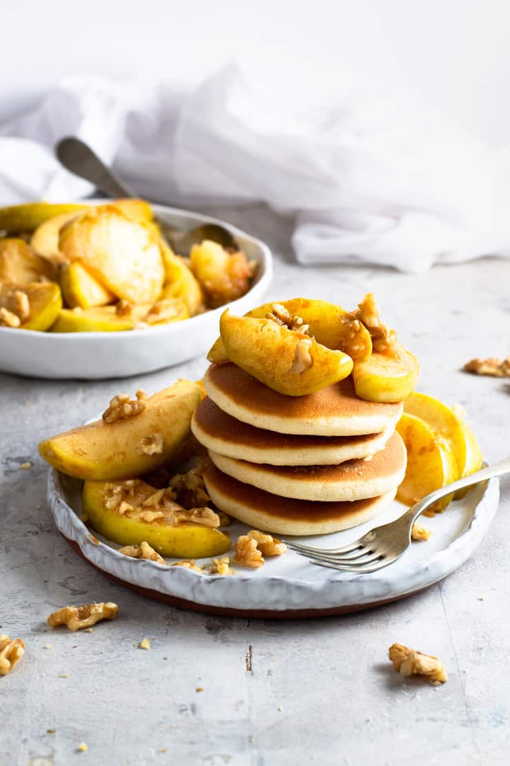 Baked apples and walnuts on top of pancakes on a small plate with a large bowl of baked apples in the background