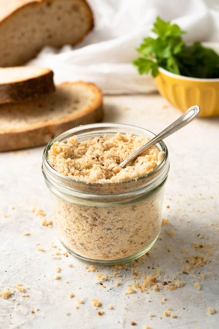 Homemade panko crumbs in a glass jar with a spoon and slices of bread in the background