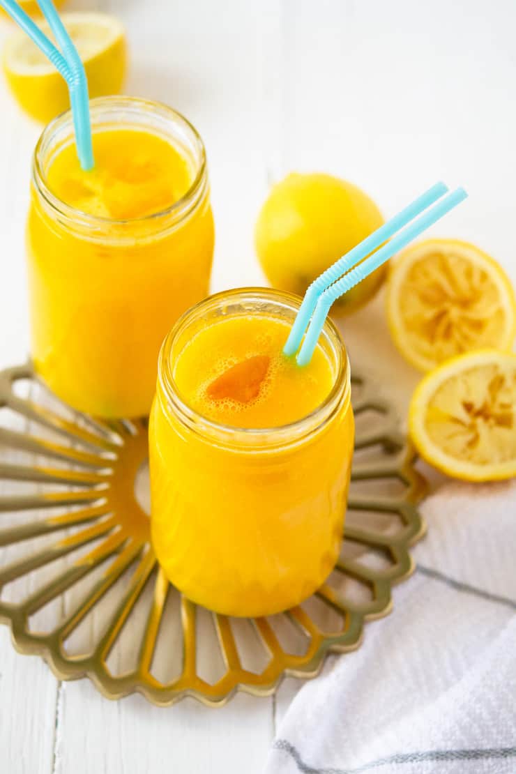 front angle view of two glasses of all natural mango lemonade with teal straws, on gold tray with white background