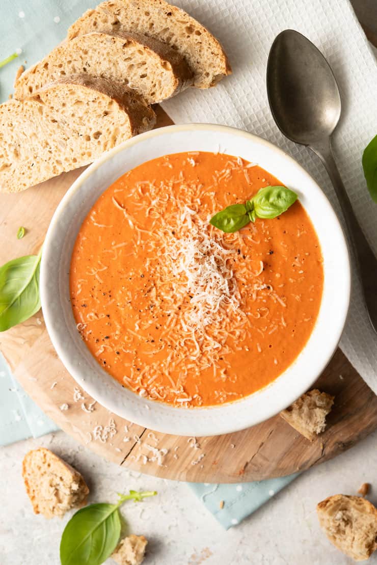 An overhead shot of roasted red pepper soup in a bowl with slices of bread and basil