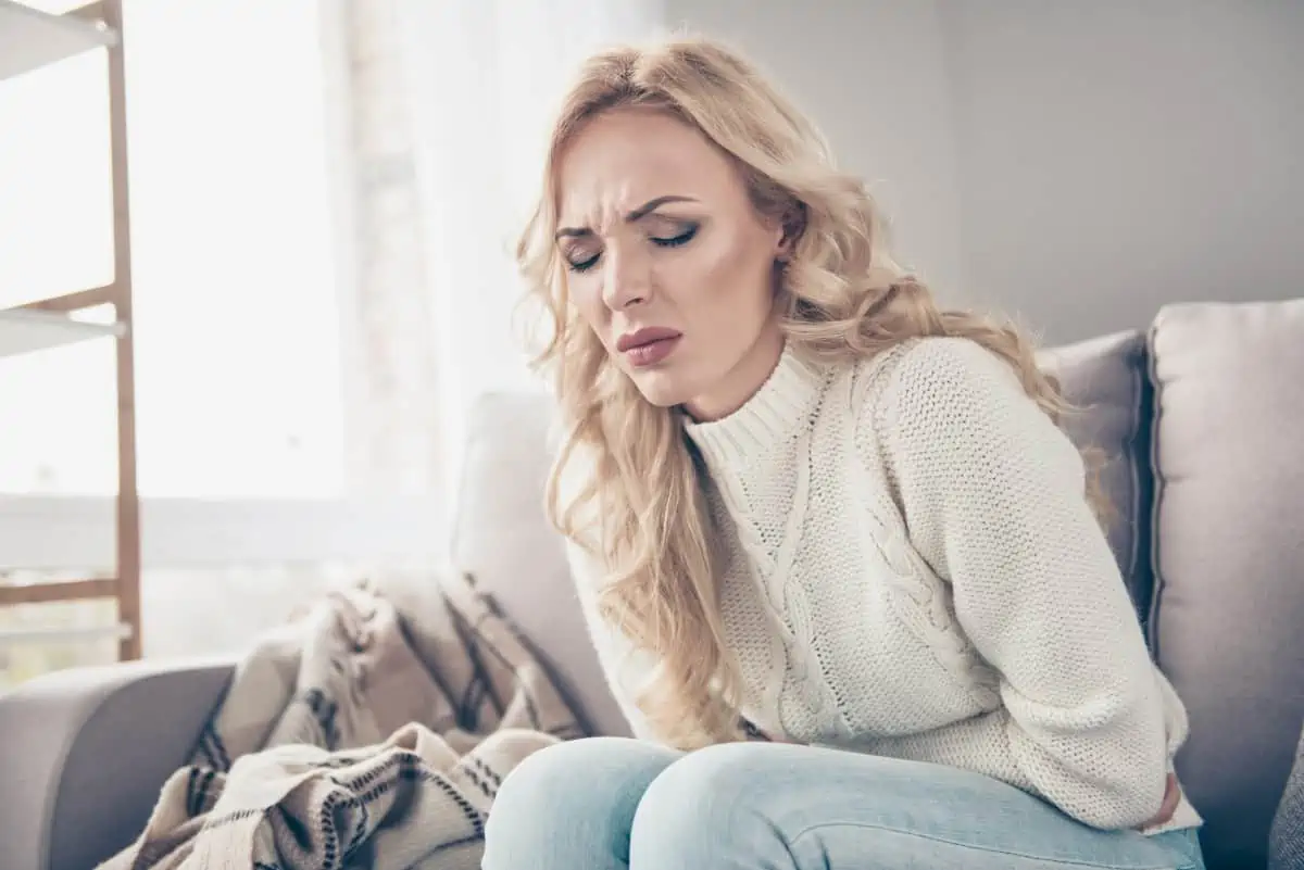 Blonde woman sitting on couch holding her stomach in pain