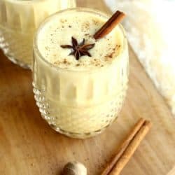 Two glasses of eggnog on wooden tray with cinnamon sticks and ground spices