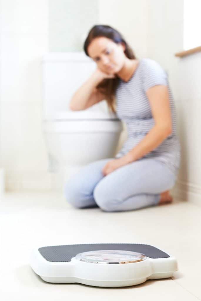 Sad young woman sitting on bathroom floor near scale