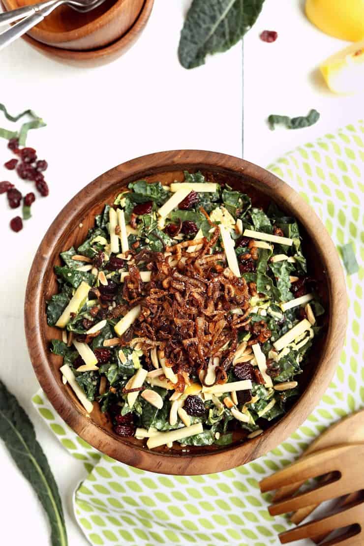 Overhead image of Kale and Brussels Sprouts Super Salad with apples, cherries, almonds, and fried shallots in wooden bowl on white background with extra ingredients sprinkled around