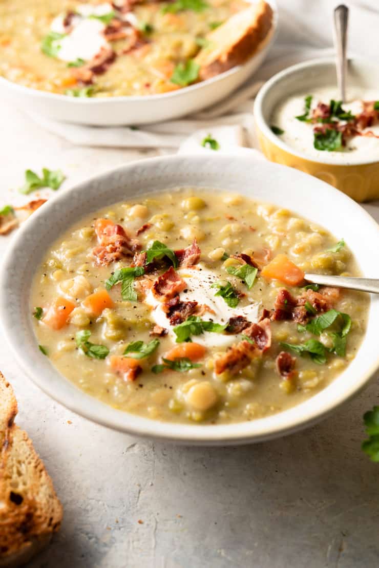 A side shot of split pea soup in a white bowl with soup cream, bacon and parsley