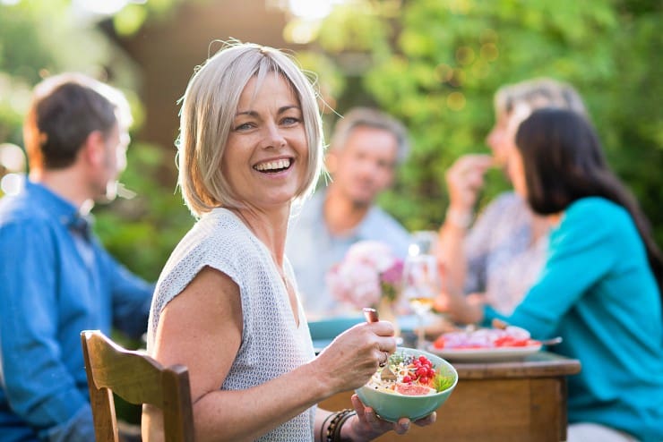 Woman at outdoor dinner party turned toward camera, smiling and holding a bowl of healthy food