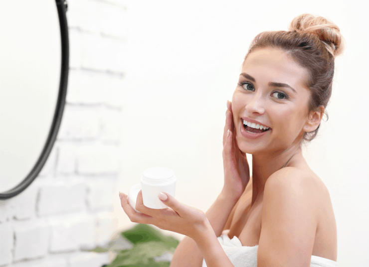 Pretty young woman applying face cream in front of bathroom mirror