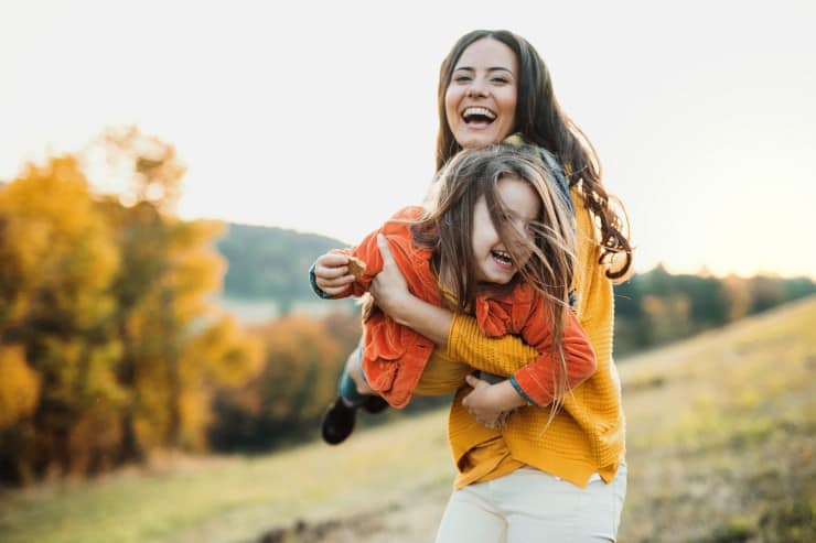 brunette woman laughing and swinging her young daughter around in a field with mountains and lake in the background