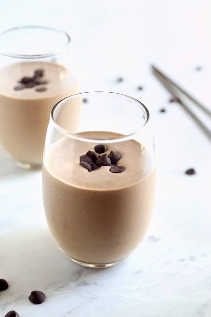 Chocolate protein shake in 2 glasses with chocolate chips on top and sprinkled around marble countertop