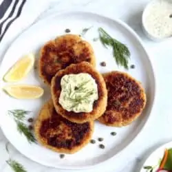 overhead image of 4 salmon burgers on white plate with tartar sauce and lemon wedges