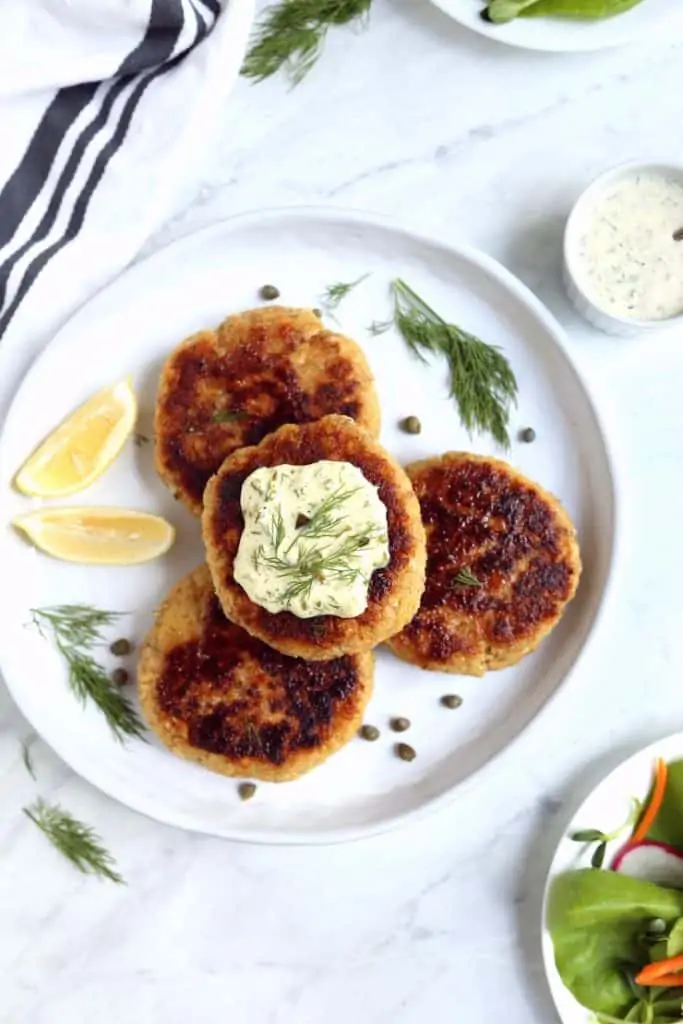 overhead image of 4 salmon burgers on white plate with tartar sauce and lemon wedges