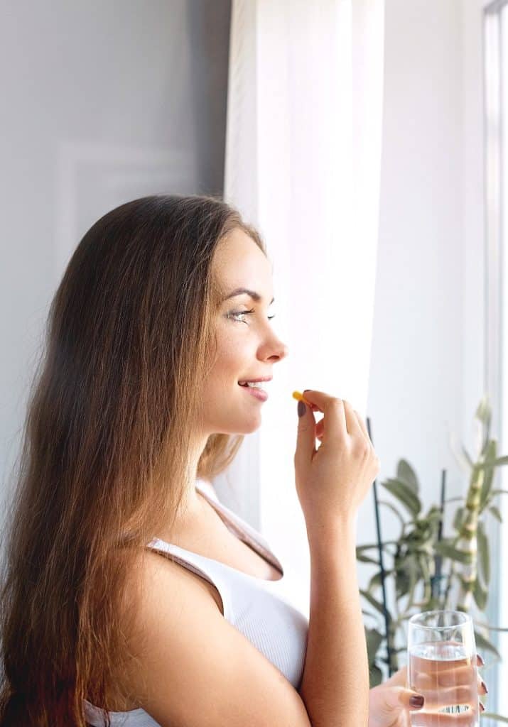 Happy young woman looking out apartment window, taking dietary supplements
