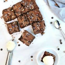 top view of batch of fudgy flourless brownies cut into squares on marble background. One square on white plate topped with ice cream.