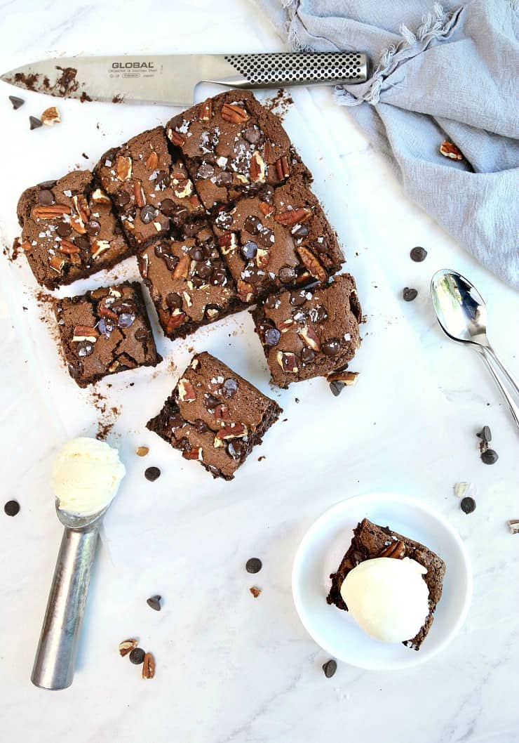 top view of batch of fudgy flourless brownies cut into squares on marble background. One square on white plate topped with ice cream.