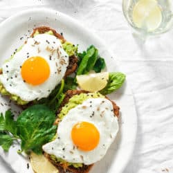 overhead view of 2 fried eggs on avocado toast with wilted greens. white plate on white background.