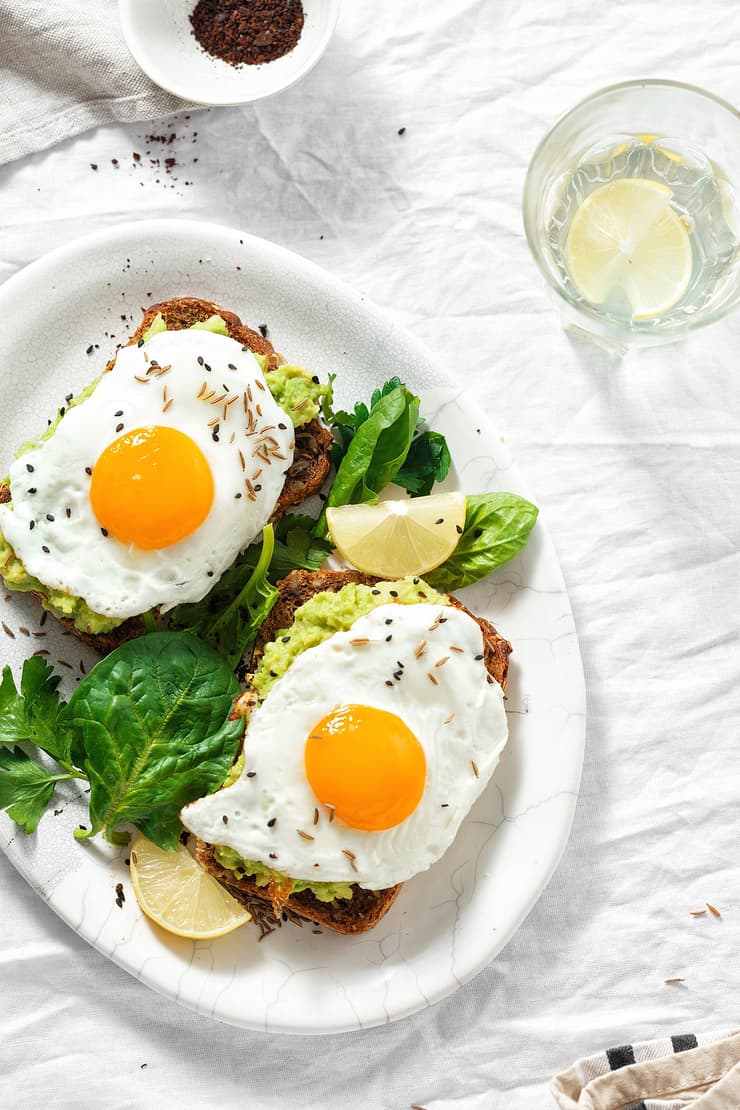 overhead view of 2 fried eggs on avocado toast with wilted greens. white plate on white background.