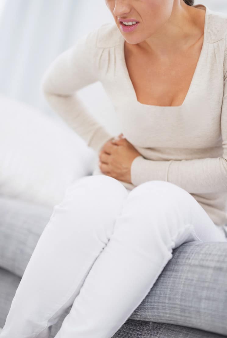 Close up image of woman seated on couch,holding her stomach in pain from Celiac Disease