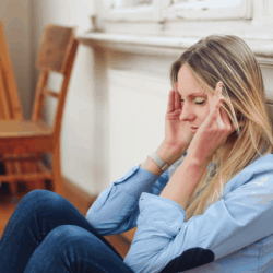 Blonde woman with migraine holding her head and sitting on kitchen floor