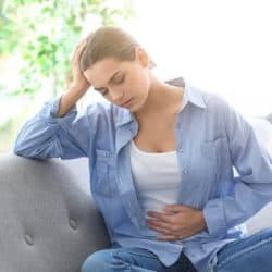 Brunette woman sitting on gray couch holding her stomach in pain