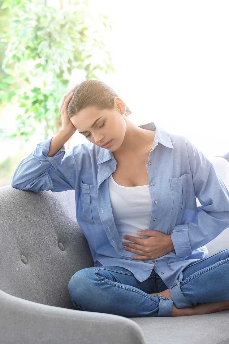 Brunette woman sitting on gray couch holding her stomach in pain