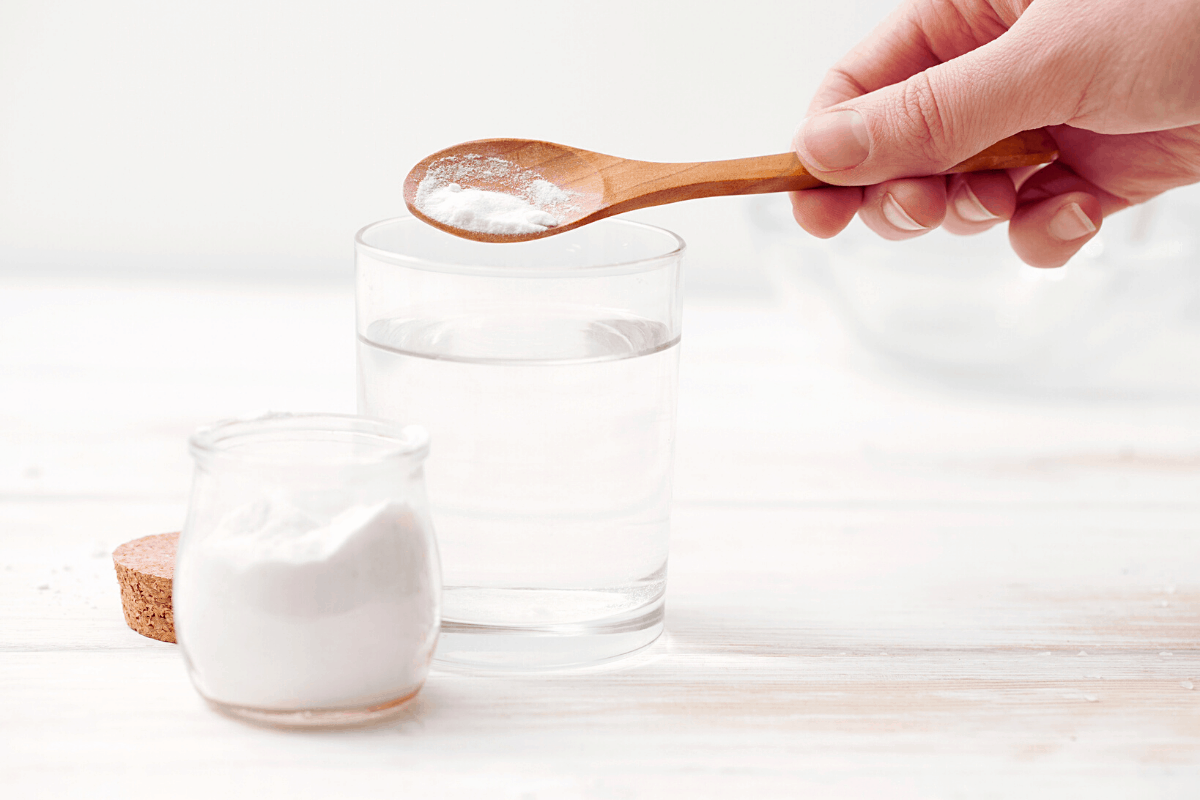 close up image of hand using wooden spoon to scoop baking soda into a glass of water 
