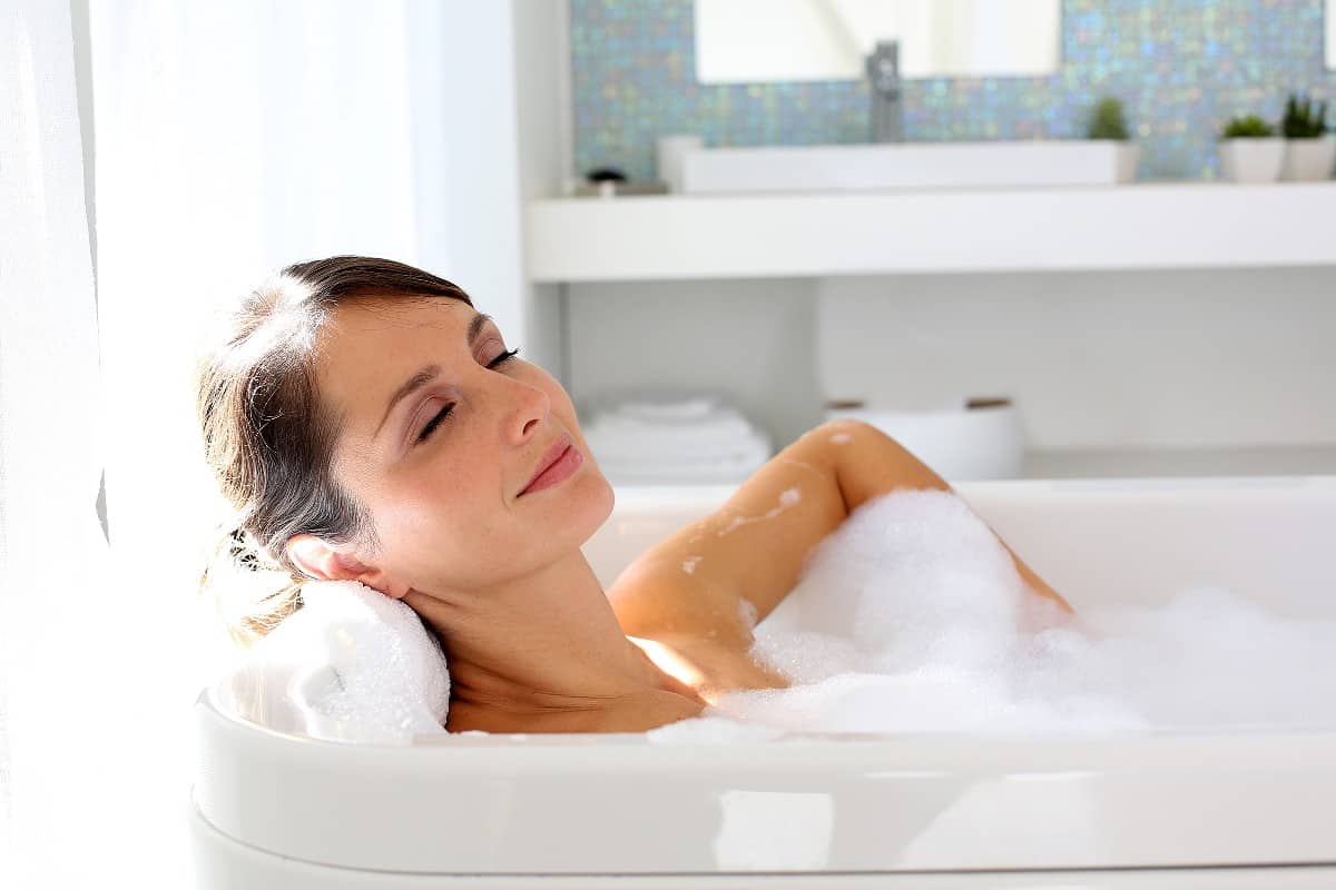 Brunette woman relaxing with her eyes closed in a bubble bath in a white tub.