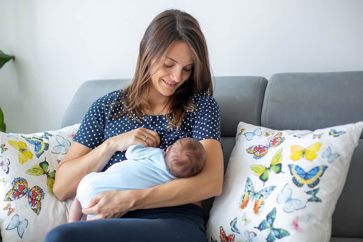 Young mom on gray couch smiling while breastfeeding her infant