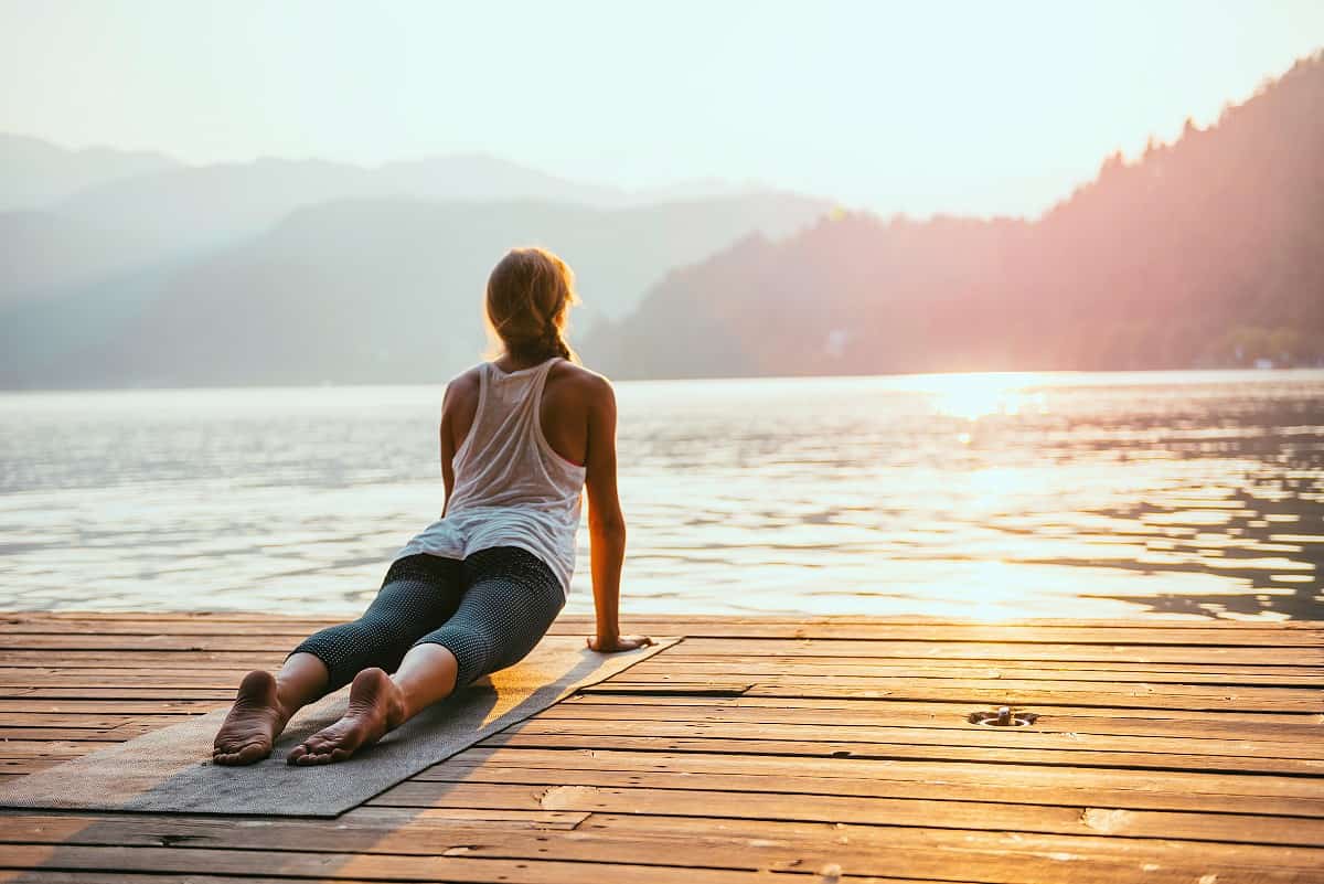 brunette woman stretching on yoga mat in front of a lake at sunrise