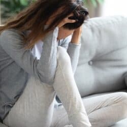 Young brunette woman on gray couch feeling anxious with head in her hands