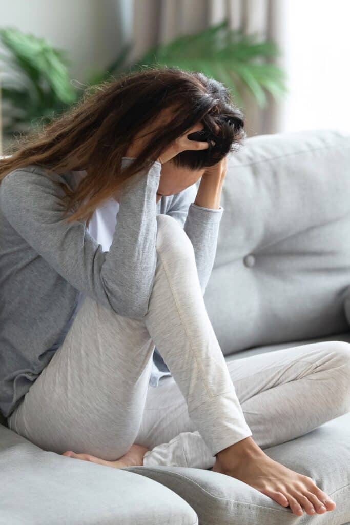 Young brunette woman on gray couch feeling anxious with head in her hands