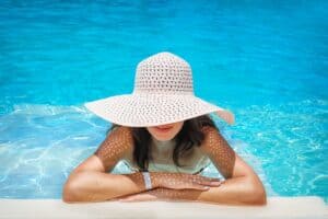 tan woman in pool, leaning arms on edge and wearing a straw sun hat