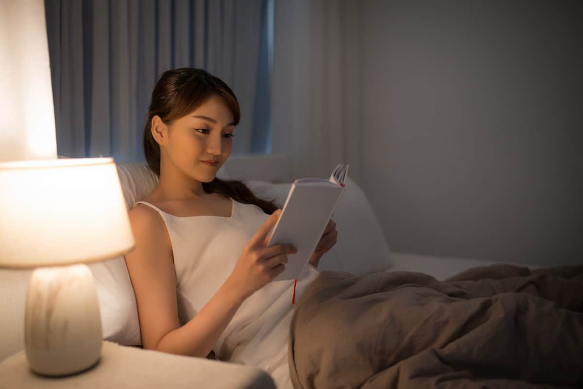 Young Asian woman reading book in bed by lamplight
