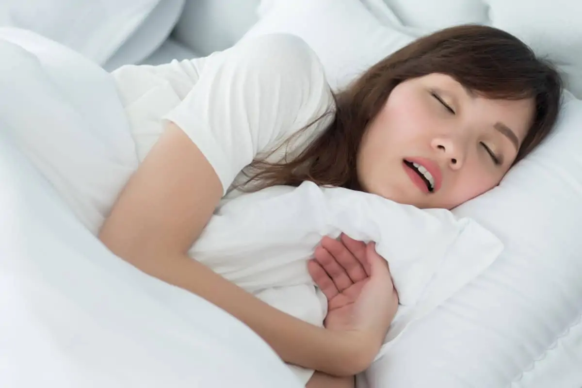 Asian woman asleep with mouth open, lying in bed with white bedding